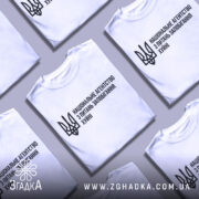 Футболка національне агентство, біла, розкладена на поверхні, фон світло-сірий, кілька футболок розташовані поруч, принт чорного кольору з текстом та тризубом.