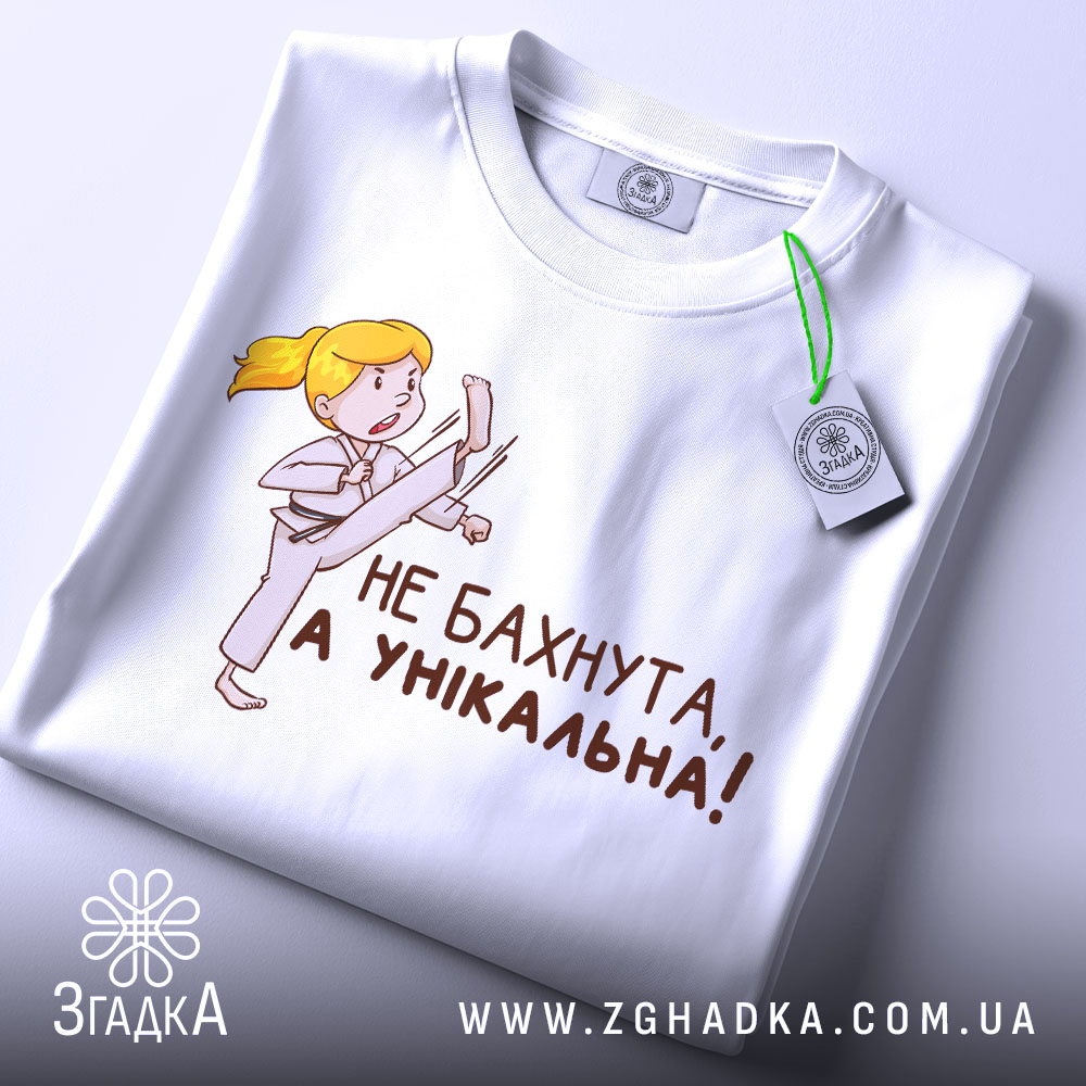 Футболка не бахнута унікальна, білого кольору, складена на поверхні, фон світло-сірий, принт з дівчинкою у кімоно, яка виконує удар, текст чорного кольору: 'НЕ БАХНУТА, А УНІКАЛЬНА!', на комірці видно етикетку.