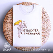 Футболка не бахнута унікальна, білого кольору, складена на плетеній кошику, фон світло-сірий, принт з дівчинкою у кімоно, яка виконує удар, текст чорного кольору: 'НЕ БАХНУТА, А УНІКАЛЬНА!'.