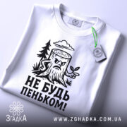 Футболка не будь пеньком. Біла футболка з чорним принтом, зображення пенька з гілочкою та написом розташоване на грудях. Вид зверху, на футболці є етикетка з зеленим шнурком. Фон світлий.