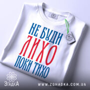 Футболка не буди лихо білого кольору, зображена зверху, складена на поверхні, фон світло-сірий, принт червоного та синього кольорів з текстом 'НЕ БУДИ ЛИХО ПОКИ ТИХО', на комірці є етикетка.