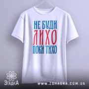 Футболка не буди лихо білого кольору, зображена на вішалці, фон світло-сірий, принт червоного та синього кольорів з текстом 'НЕ БУДИ ЛИХО ПОКИ ТИХО'.