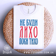 Футболка не буди лихо білого кольору, зображена зверху, складена на плетеній підставці, фон світло-сірий, принт червоного та синього кольорів з текстом 'НЕ БУДИ ЛИХО ПОКИ ТИХО'.