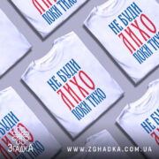 Футболка не буди лихо білого кольору, зображена з фронтального ракурсу на манекені, фон світло-сірий, принт червоного та синього кольорів з текстом 'НЕ БУДИ ЛИХО ПОКИ ТИХО'.