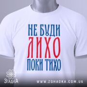 Футболка не буди лихо білого кольору, зображена зверху, складена на поверхні, фон світло-сірий, принт червоного та синього кольорів з текстом 'НЕ БУДИ ЛИХО ПОКИ ТИХО', на комірці є етикетка.