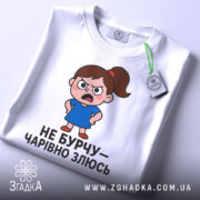 Футболка не бурчу. Біла футболка, зображена з верхнього ракурсу, на ній малюнок сердитої дівчинки в синій сукні, текст чорного кольору, на комірці видно етикетку.
