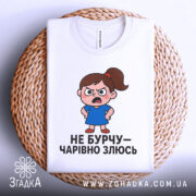 Футболка не бурчу. Біла футболка, складена на плетеній основі, з малюнком сердитої дівчинки в синій сукні, текст чорного кольору, фон світлий.