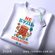 Футболка не бути пеньком. Біла футболка, складена на поверхні, з кольоровим принтом пенька, який усміхається і тримає гриб. На футболці є етикетка з зеленим шнурком. Ракурс зверху.