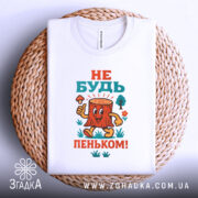 Футболка не бути пеньком. Біла футболка, складена на плетеній основі, з кольоровим принтом пенька, який усміхається і тримає гриб. Ракурс зверху, фон світлий.