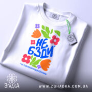 Футболка не бзди, біла, складена на поверхні, фон світло-сірий, принт з яскравими квітами та текстом "НЕ БЗДИ" у синьому кольорі, з етикеткою на зеленому шнурку.