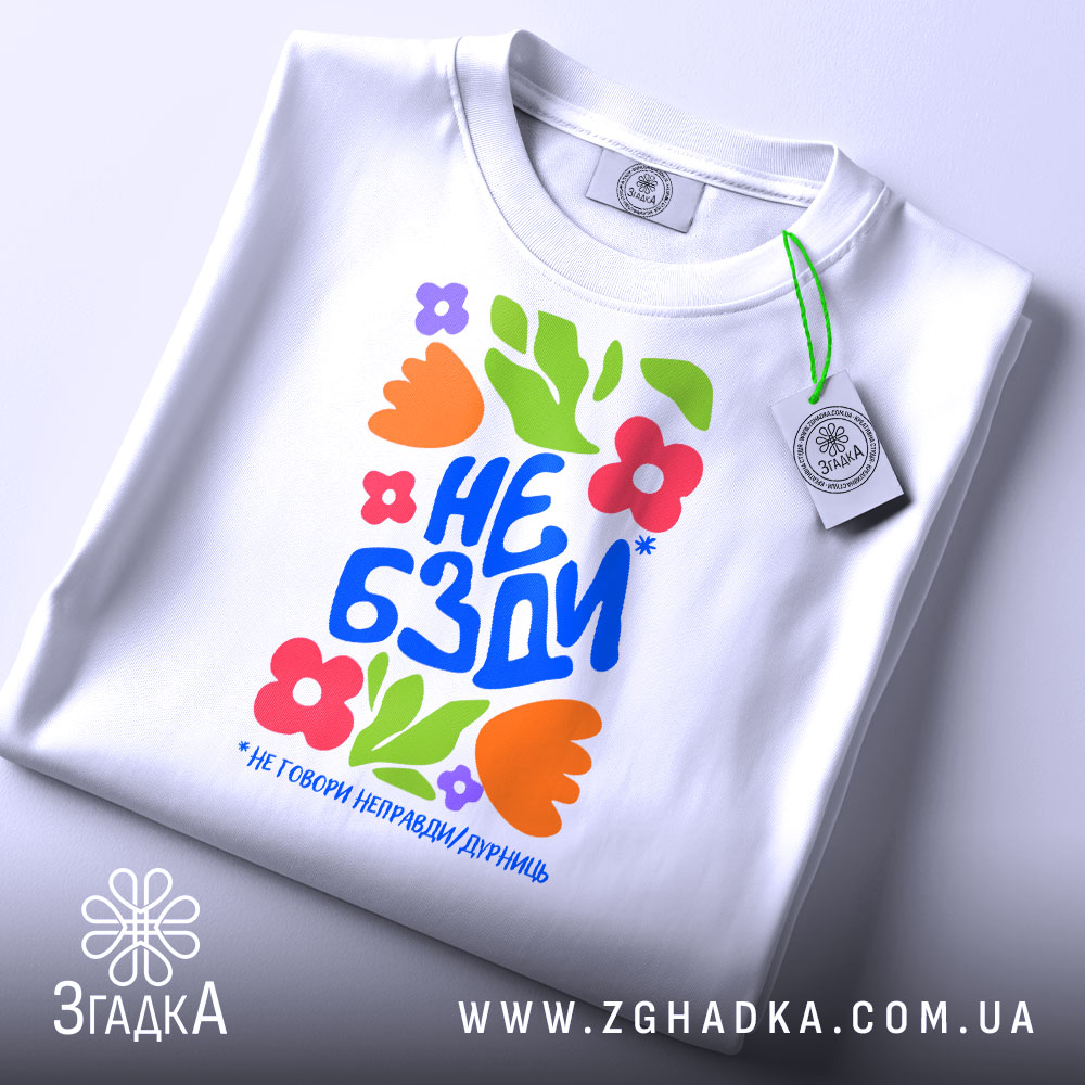 Футболка не бзди, біла, складена на поверхні, фон світло-сірий, принт з яскравими квітами та текстом "НЕ БЗДИ" у синьому кольорі, з етикеткою на зеленому шнурку.