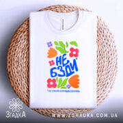 Футболка не бзди, біла, складена на поверхні з плетеного матеріалу, фон світло-сірий, принт з яскравими квітами та текстом "НЕ БЗДИ" у синьому кольорі.