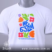 Футболка не бзди, біла, складена на поверхні, фон світло-сірий, принт з яскравими квітами та текстом "НЕ БЗДИ" у синьому кольорі, з етикеткою на зеленому шнурку.