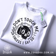 Футболка не чіпай мене. Біла футболка, складена на столі, з принтом чорного черепа з рогами, фон світло-сірий, на футболці є етикетка з зеленим шнурком, принт з написом 'DON'T TOUCH ME UNLESS I SAY SO'.