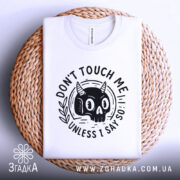 Футболка не чіпай мене. Біла футболка, складена на плетеному кошику, фон світло-сірий, принт з чорним черепом з рогами та написом 'DON'T TOUCH ME UNLESS I SAY SO'.