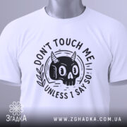 Футболка не чіпай мене. Біла футболка, складена на столі, з принтом чорного черепа з рогами, фон світло-сірий, на футболці є етикетка з зеленим шнурком, принт з написом 'DON'T TOUCH ME UNLESS I SAY SO'.