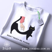 Футболка не чіпай. Складена біла футболка з принтом чорного кота та руки, що його торкається. На футболці є етикетка з зеленим шнурком. Фон світло-сірий.