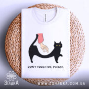 Футболка не чіпай. Складена біла футболка на плетеній кошику, з принтом чорного кота та руки, що його торкається. Фон світло-сірий, принт яскравий, текст чорний.