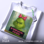 Футболка не чіпайте бантик. Біла футболка, на якій зображений зелений персонаж з рожевим бантиком. Вид з верхнього ракурсу, на футболці є етикетка з зеленим шнурком. Фон світлий, з легким відблиском.