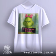 Футболка не чіпайте бантик. Біла футболка з принтом, на якому зелений персонаж з рожевим бантиком. Фон однорідний, з легким відтінком сірого. Текст на принті чорного кольору, розташований внизу зображення.