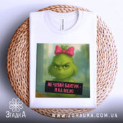 Футболка не чіпайте бантик. Біла футболка, складена на плетеній основі. Принт з зеленим персонажем з рожевим бантиком, фон розмитий, з відтінками зеленого. Текст на принті чорного кольору.