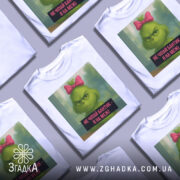 Футболка не чіпайте бантик. Біла футболка з принтом, на якому зображений зелений персонаж з рожевим бантиком на голові. Фон розмитий, з відтінками зеленого та блакитного. Текст на принті чорного кольору з білим шрифтом.