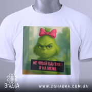 Футболка не чіпайте бантик. Біла футболка, на якій зображений зелений персонаж з рожевим бантиком. Вид з верхнього ракурсу, на футболці є етикетка з зеленим шнурком. Фон світлий, з легким відблиском.