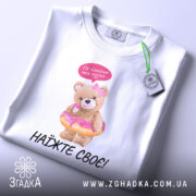 Футболка не чіпайте пузіко. Біла футболка, зображена з верхнього ракурсу, ведмедик у купальнику з пончиком, на фоні світло-сірого градієнту. Принт яскравий, ведмедик тримає пончик, зверху напис "Не чіпайте моє пузіко...". На футболці є етикетка.