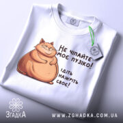 Футболка не чіпайте пузіко. Біла футболка, складена на поверхні, з принтом кота та текстом. Видно етикетку з зеленим шнурком, фон світлий.
