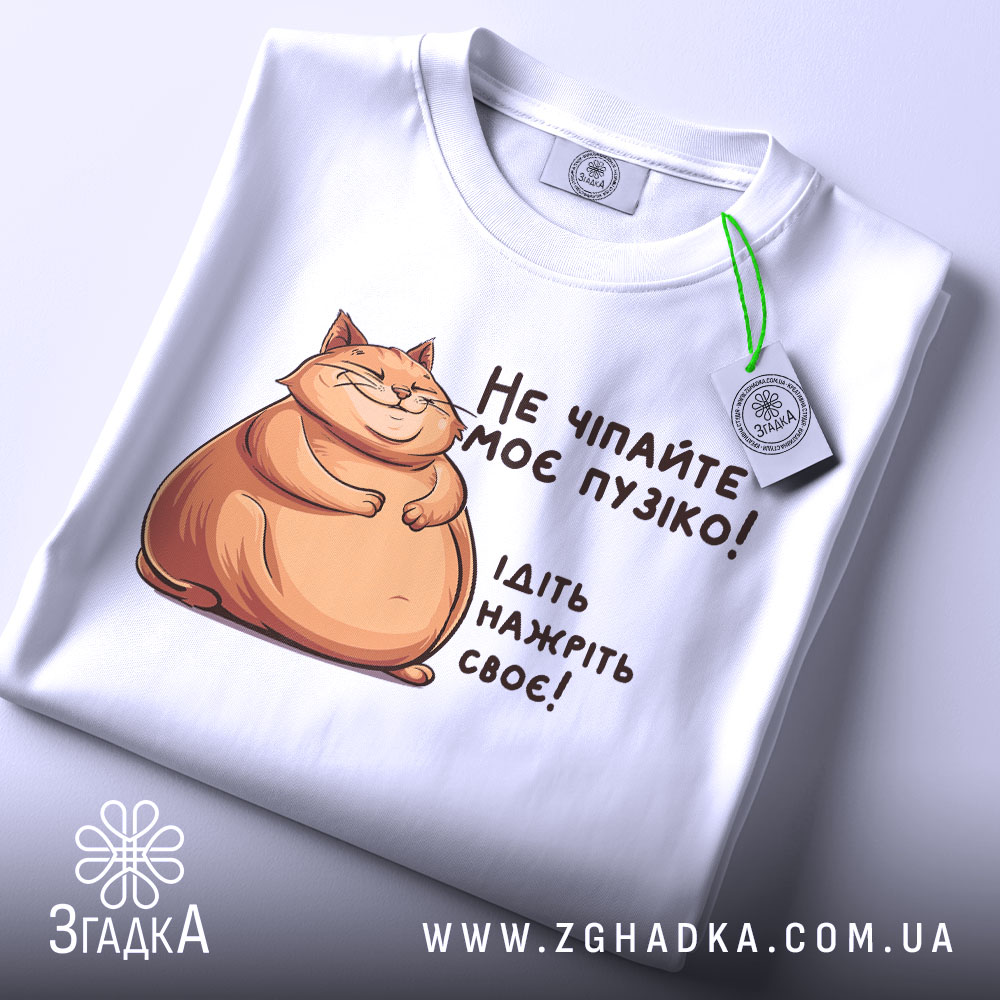 Футболка не чіпайте пузіко. Біла футболка, складена на поверхні, з принтом кота та текстом. Видно етикетку з зеленим шнурком, фон світлий.