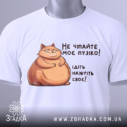 Футболка не чіпайте пузіко. Біла футболка, складена на поверхні, з принтом кота та текстом. Видно етикетку з зеленим шнурком, фон світлий.