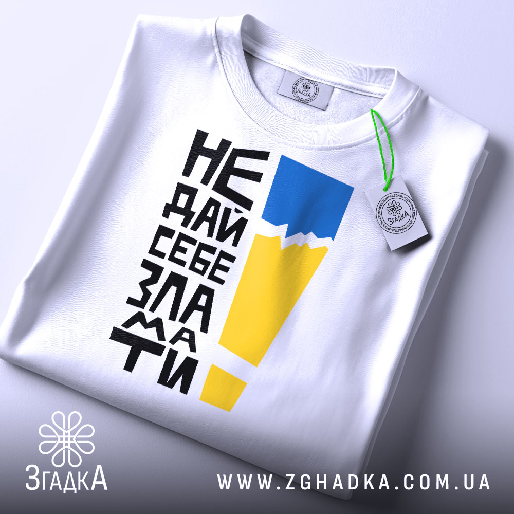 Футболка не дай себе зламати. Біла футболка, складена на плоскій поверхні, з видимим етикеткою. На фоні світло-сірого кольору. Принт з чорного тексту, жовтого і блакитного кольорів, розташований вертикально.