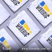 Футболка не дай себе зламати. Біла футболка з коротким рукавом, зображена з правого ракурсу. На фоні світло-сірого кольору. Принт містить чорний текст з жовтим і блакитним елементами, що створює контрастний вигляд.