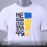 Футболка не дай себе зламати. Біла футболка, складена на плоскій поверхні, з видимим етикеткою. На фоні світло-сірого кольору. Принт з чорного тексту, жовтого і блакитного кольорів, розташований вертикально.
