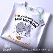 Футболка не довбіть мозок, біла, складена на поверхні, фон світлий, принт з малюнком мозку та текстом "Не добийте мені МІЙ МОЗОК ВІН У ВІДПУСТЦІ!!" у чорному та оранжевому кольорах, з етикеткою зеленого кольору.