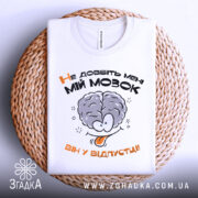 Футболка не довбіть мозок, біла, складена на плетеній основі, фон світлий, принт з малюнком мозку та текстом "Не добийте мені МІЙ МОЗОК ВІН У ВІДПУСТЦІ!!" у чорному та оранжевому кольорах.