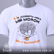 Футболка не довбіть мозок, біла, складена на поверхні, фон світлий, принт з малюнком мозку та текстом "Не добийте мені МІЙ МОЗОК ВІН У ВІДПУСТЦІ!!" у чорному та оранжевому кольорах, з етикеткою зеленого кольору.