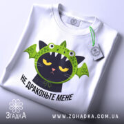 Футболка не драконьте мене, біла, складена на столі, фон світлий, принт з чорним котом у зеленій драконячій шапці, з етикеткою на зеленій стрічці.