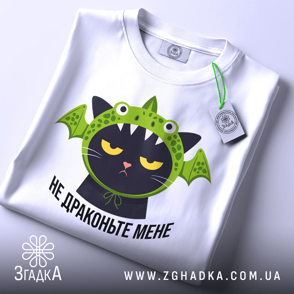 Футболка не драконьте мене, біла, складена на столі, фон світлий, принт з чорним котом у зеленій драконячій шапці, з етикеткою на зеленій стрічці.