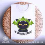 Футболка не драконьте мене, біла, складена на плетеному кошику, фон світлий, принт з чорним котом у зеленій драконячій шапці, з виразом незадоволення.