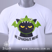 Футболка не драконьте мене, біла, складена на столі, фон світлий, принт з чорним котом у зеленій драконячій шапці, з етикеткою на зеленій стрічці.