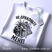 Футболка не драконьте мене. Біла футболка, складена на плоскій поверхні, з видимим принтом чорного дракона та текстом "НЕ ДРАКОНЬТЕ МЕНЕ!". На футболці є етикетка з зеленим шнурком, фон світло-сірий.