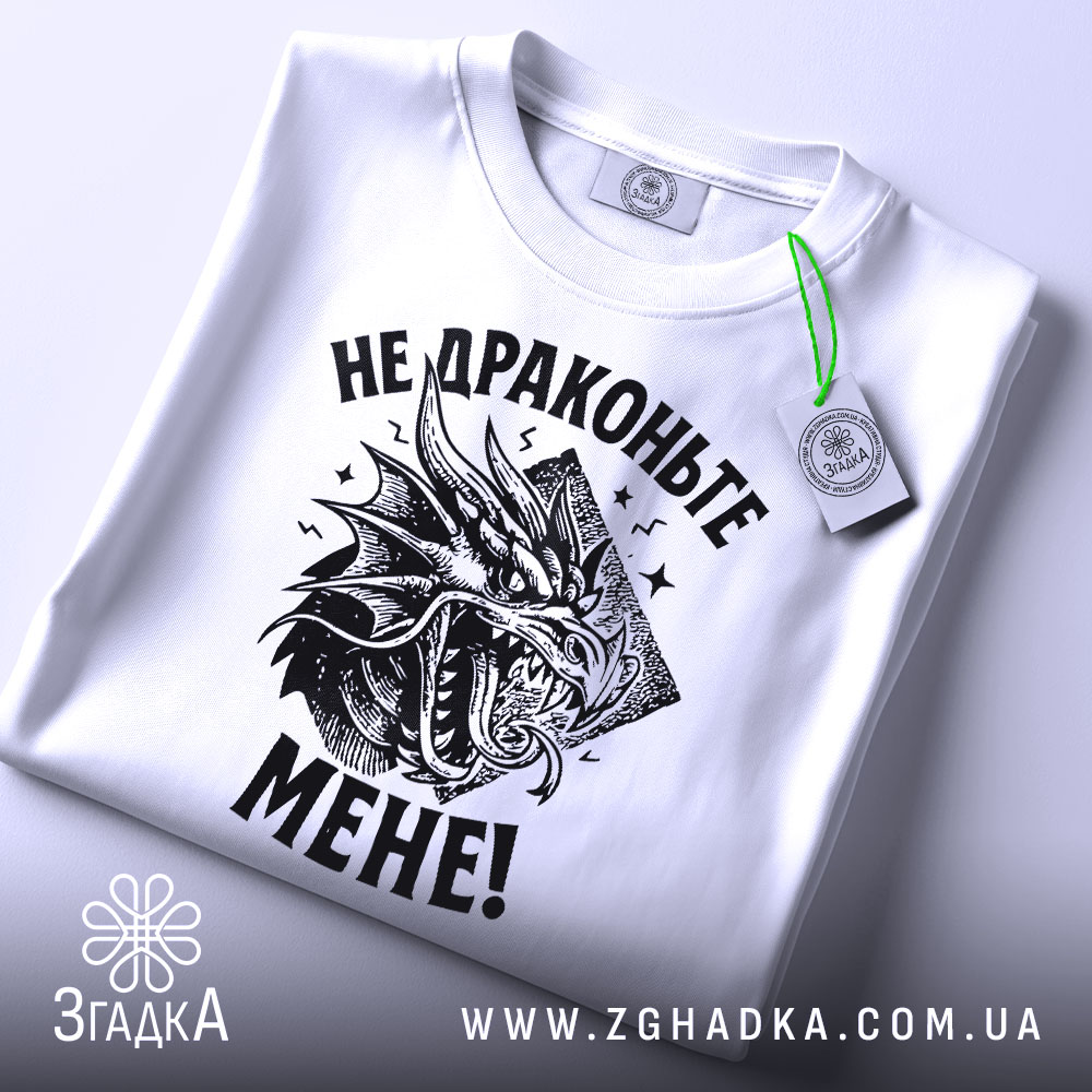 Футболка не драконьте мене. Біла футболка, складена на плоскій поверхні, з видимим принтом чорного дракона та текстом "НЕ ДРАКОНЬТЕ МЕНЕ!". На футболці є етикетка з зеленим шнурком, фон світло-сірий.