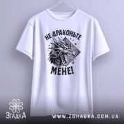 Футболка не драконьте мене. Біла футболка з коротким рукавом, підвішена на вішалці, з принтом чорного дракона та текстом "НЕ ДРАКОНЬТЕ МЕНЕ!" на грудях. Фон світло-сірий.