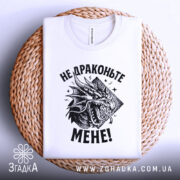 Футболка не драконьте мене. Біла футболка, складена на плоскій поверхні з плетеного кошика, з принтом чорного дракона та текстом "НЕ ДРАКОНЬТЕ МЕНЕ!". Фон світло-сірий.