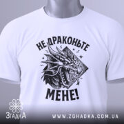 Футболка не драконьте мене. Біла футболка, складена на плоскій поверхні, з видимим принтом чорного дракона та текстом "НЕ ДРАКОНЬТЕ МЕНЕ!". На футболці є етикетка з зеленим шнурком, фон світло-сірий.