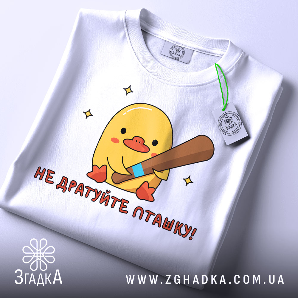 Футболка не дратуйте пташку, біла, складена на столі, фон світло-сірий, принт з жовтим каченям, яке тримає биту, текст червоним шрифтом, на комірці видна етикетка.