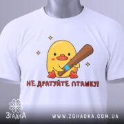 Футболка не дратуйте пташку, біла, складена на столі, фон світло-сірий, принт з жовтим каченям, яке тримає биту, текст червоним шрифтом, на комірці видна етикетка.