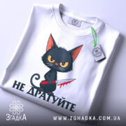 Футболка не дратуйте, білого кольору, зображена зверху, на фоні світло-сірого кольору, принт з чорним котом з червоним ножем, текст під котом чорного кольору, на футболці є етикетка з зеленим шнурком.