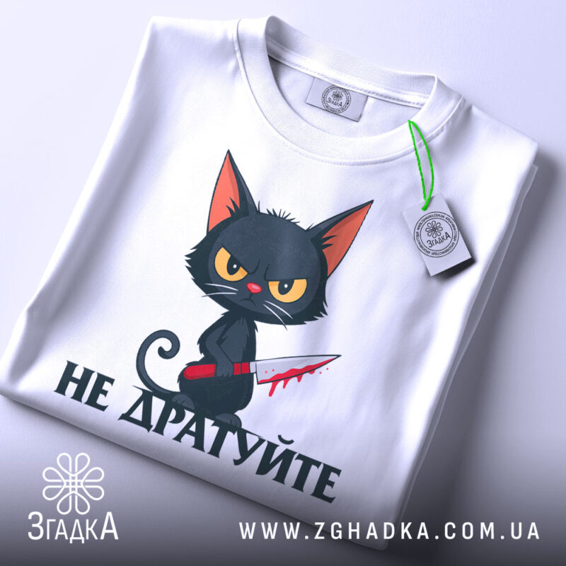 Футболка не дратуйте, білого кольору, зображена зверху, на фоні світло-сірого кольору, принт з чорним котом з червоним ножем, текст під котом чорного кольору, на футболці є етикетка з зеленим шнурком.